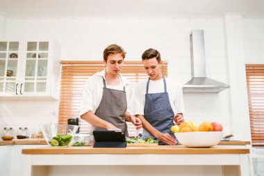 Önlük giyen iki genç adam, tabletteki tarifi takip ederek, modern bir mutfakta taze bir salata hazırlıyorlar. Sahne, sağlıklı beslenmeyi, birlikte yemek yapmayı ve evde dengeli bir yaşam tarzının keyfini çıkarmayı ön plana çıkarıyor.