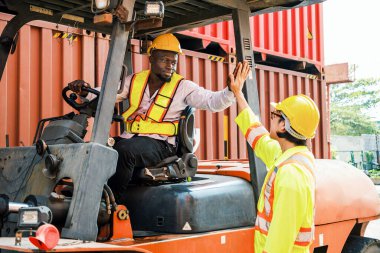Güvenlik teçhizatındaki iki depo çalışanı bir nakliye deposunda beşlik çakıyor. Bir forklift operatörü ve bir meslektaşı yığınla kargo konteynırı arasında takım çalışmasını ve işbirliğini kutladılar.