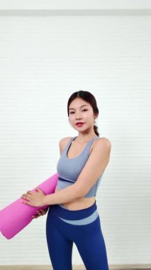 Genç bir kadın, spor kıyafeti giymiş pembe bir yoga minderi tutuyor. İyi aydınlatılmış bir yerde antrenmana hazır. Spor kıyafetleri giyerek evde spor yapıyor ya da egzersiz yapıyor..