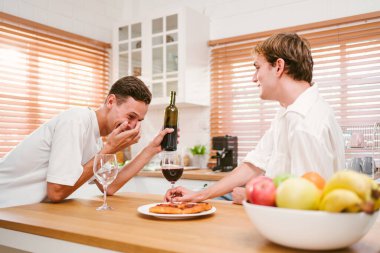 İki genç adam modern mutfakta şarap ve pizzanın tadını çıkarırken aynı kahkahayı paylaşıyor. Sıcak ortam arkadaşlığı, bağı, rahatlamayı, gelişigüzel yemek yemeyi, mutluluğu ve sosyal bağlantıyı temsil ediyor..