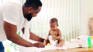 Sevgi dolu bir Kara Baba, bebek bezi değiştirirken güç şişesini ısıran bebeğine bakar. Ebeveyn olmak, kaynaşmak ve doğal ışıkla sıcak bir evde babalık anları. Gerçek zamanlı 4k video