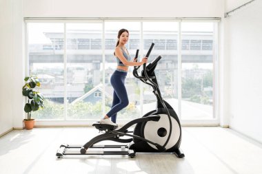 Parlak bir spor salonunda eliptik bir eğitmen üzerinde egzersiz yapan bir kadın. Kardiyo, kilo kaybı, fitness rutini, sağlıklı yaşam tarzı, kapalı alanda egzersiz, kişisel eğitim ve ev egzersizi gibi kavramlar için mükemmel..