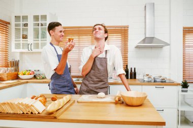 Önlüklü iki neşeli LGBT fırıncısı sıcak bir mutfakta taze ekmek hazırlarken kahkahalarını paylaşıyorlar. Takım çalışmaları ve tutkuları aşçılık ve aşçılık sanatlarında aşkı, kapsayıcılığı ve yaratıcılığı yansıtır..