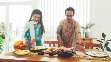 Geleneksel Hint kıyafetli çift evde şenlikli bir yemek yiyor. Masa lezzetli Hint yemekleriyle dolu. Kültürü, kutlamayı, özgün ev yemeklerini sembolize ediyor. Yavaş çekim 4k video.