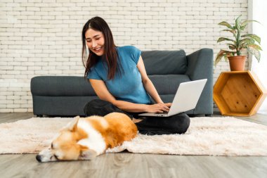 Corgi köpeği rahat bir halıda uyurken genç bir kadın dizüstü bilgisayarıyla çalışıyor. Sahne, uzaktan çalışma, evcil hayvan arkadaşlığı, iş hayatı dengesi ve rahat bir ev ortamı yakalıyor..