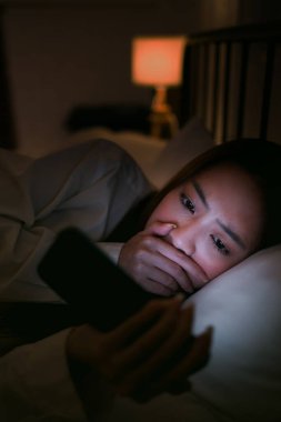 Genç bir kadın gece yatakta, akıllı telefonuna bakarken şok olmuş ve endişeli. Dijital stres, sosyal medya kaygısı, siber zorbalık, kötü haber ve Gen Z üzerindeki zihinsel sağlık etkileri kavramı.