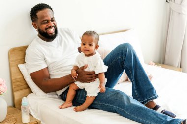 Mutlu Afrikalı baba ve bebek evde rahat bir yatakta kaynaşıyor. Sevgi, neşe ve kahkahayla dolu, sevgi dolu bir ebeveyn anı. Gösterişli bir baba, aile bağı ve şefkatli bir bakım..