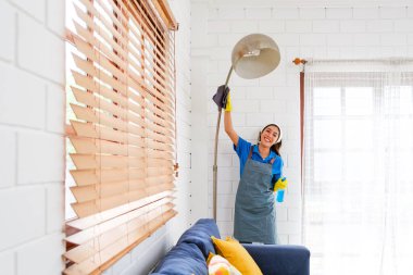 Gülümseyen Asyalı kadın üniformalı, parlak oturma odasında lambanın tozunu alıyor. Temizlik, ev temizliği ve düzenli bir iç mekan için hijyen bakımı..