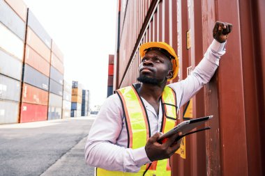 Sıkı bir şapka ve güvenlik yeleği giymiş Afrikalı bir lojistik işçisi bir kargo limanında elinde tablet olan konteynerleri denetliyor. Yük yönetimi, tedarik zinciri lojistiği ve küresel ticaret operasyonları iş başında