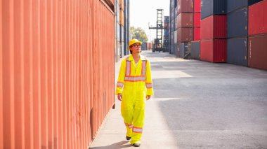 Yüksek görüş açısına sahip güvenlik giysisiyle Asyalı bir lojistik işçisi istiflenmiş kargo konteynırlarıyla çevrili bir nakliye sahasında yürüyor. Yük taşımacılığı, küresel ticaret, tedarik zinciri yönetimi.
