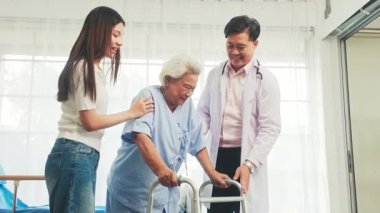 Doktor ve bakıcı yaşlı bir kadına rehabilitasyon merkezinde yürüteç kullanmasında yardım ediyor. Yaşlı bakımı, fizyoterapi, taşınabilirlik desteği, bir sağlık merkezinde hasta tedavisi. 4k gerçek zamanlı video.