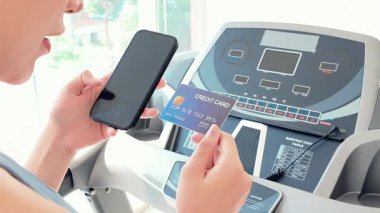 Kadının elinde akıllı telefon ve kredi kartı var. Koşu bandının yanında duruyor. Muhtemelen online ödeme ya da abonelik yapıyor. Dijital işlemler ve zindelik alımları. Yavaş çekim 4k video