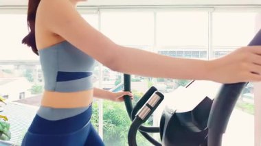Şık, aktif giyinen genç bir kadın, parlak bir spor salonunda eliptik bir makinede egzersiz yapıyor. Y jenerasyonu ve Z jenerasyonu sağlık, kardiyo ve kişisel bakım sağlığına odaklı yaşam tarzı. Yavaş çekim 4k video