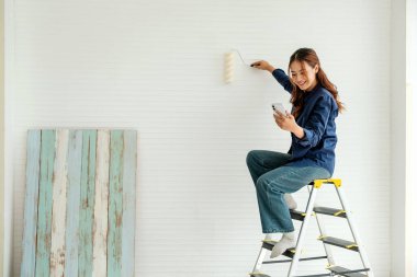 Duvarı silindirle boyarken ve akıllı telefon kullanırken merdivende oturan genç bir kadın. DIY ev yenileme projesi, parlak bir alanda modern teknoloji ve yaratıcılık ile çoklu görev.