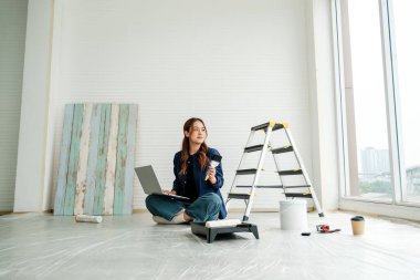 Genç bir kadın, elinde fırça ve dizüstü bilgisayar ile DIY ev yenileme projesi yapıyor. Parlak bir alanda araçlar, merdiven ve kahve ile yaratıcı iç tasarım ve ev geliştirme kavramı.
