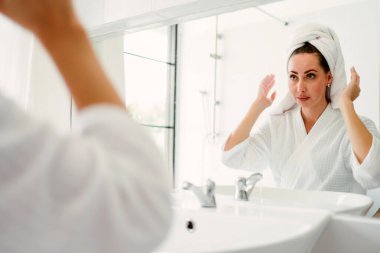 Beyaz bornoz ve havlu giymiş bir kadın sabah rutini sırasında banyo aynasında dikiliyor. Işıl ışıl, minimal ev iç mekanında özbakıma, sağlığa ve hijyene odaklanmış..