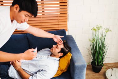 Evde oturan eşcinsel çift, hasta ortağını kanepede sevgiyle kolluyor ve ateşini kontrol ediyor. LGBTQ ilişkisi, şefkat ve günlük hayatta destekleyici sağlık konsepti.