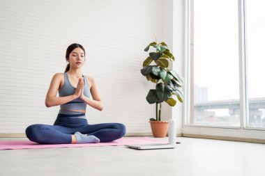 Genç Asyalı bir kadın parlak modern bir odada pembe bir paspasın üzerinde solunum ya da prana yoga meditasyonu yapıyor. Sağlık, sağlık, öz bakım ve evdeki dengeli yaşam tarzını destekliyor..