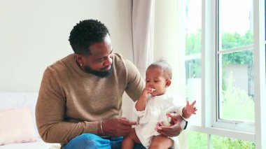 Sevgi dolu bir baba, bebeğini güneşli bir pencerenin yanında tutar ve hassas bir aile anını paylaşır. Babalar Günü, ebeveynlik ve otantik çeşitlilik için ideal bir ortam..