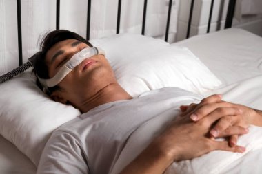 Horlama ve obstrüktif uyku apnesi (OSA) tedavisi için kullanılan CPAP burun maskesi ile huzur içinde uyuyan adam. Evde daha iyi nefes almayı, uyku sağlığını ve gece solunum terapisini teşvik eder..