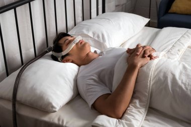 Yatakta CPAP burun maskesi takmış Asyalı bir erkek horlama ve engelleyici uyku apnesi (OSA) tedavisi görüyor. Daha iyi nefes almayı ve evde solunum terapisiyle rahat uyumayı teşvik eder..