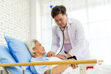 Hastane yatağında gülümseyen yaşlı bir hastayı muayene etmek için steteskop kullanan bir pratisyen hekim. Güven, sağlık ve yaşlı bakım kavramı, sağlık, sigorta ve sağlık konuları için idealdir..