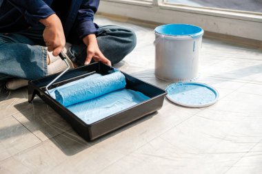 Bir kadın DIY ev yenileme projesinde çalışırken mavi boya silindiri hazırlıyor. Doğal ışık ve araçlar yaratıcılığı, ev gelişimini ve iç tasarım çabalarını ön plana çıkarıyor.