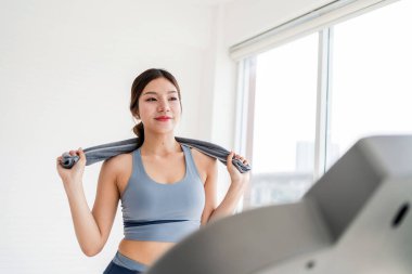 Koşu bandı antrenmanından sonra evde serinleyen genç bir kadın, omuzlarının üzerinde bir havlu tutuyor, motivasyon, sağlık ve kişisel gelişimi simgeliyor parlak, minimalist bir alanda..