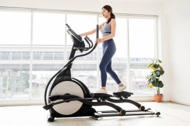 Parlak bir spor salonunda eliptik bir makine üzerinde egzersiz yapan genç bir kadın, kardiyo fitness, vücut şekillendirme ve zihinsel berraklık üzerine odaklanıyor, sağlıklı bir yaşam tarzı ve kapalı alanda çalışma motivasyonunu teşvik ediyor..