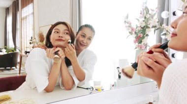 Beyaz cüppeli anne ve kız makyaj yaparken makyaj yapıyor. Gösterişli bir aynanın önünde gülümsüyor, özel bir Anneler Günü anı için parlak, rahat bir mekanda kaliteli zaman ve özgüvenin tadını çıkarıyor..