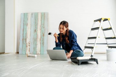 Genç bir kadın dizüstü bilgisayarında araştırma yaparken elinde boya fırçası tutuyordu. Aydınlık bir odada aletler, merdiven ve ahşap paneller ile çevrili, ev geliştirme planlama odaklı.