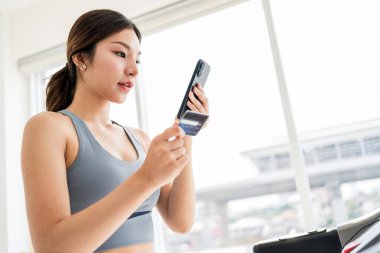 Spor kıyafeti giyen sağlıklı genç bir kadın akıllı telefon ve kredi kartı taşırken koşu bandında, refahı ve modern yaşam tarzını harmanlıyor, kolaylığı, e-ticareti ve fitness motivasyonunu simgeliyor..