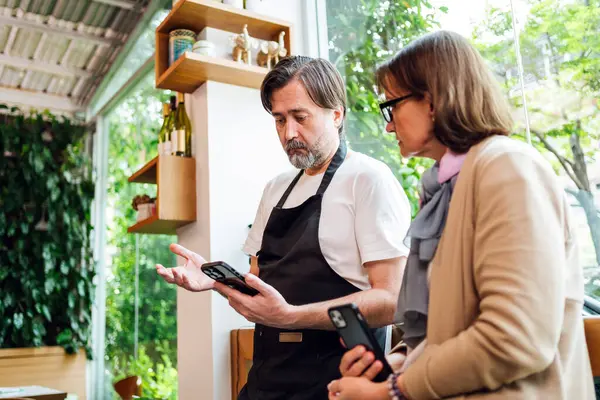 Kafe sahibi danışman ile mali stratejiyi tartışıyor, akıllı telefonlar tutuyor, rahat bir dükkanın içinde. İşbirliği, küçük işletmeler, dijital planlama, girişimcilik ve modern ekonomik büyümeyi ön plana çıkarıyor.