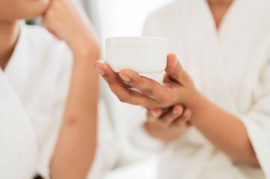 Beyaz spa bornozlu bir kadının elinde minimal beyaz kozmetik bir kavanoz, cilt bakımı, sağlık ve günlük güzellik ritüelleri, özbakım, spa ve kozmetik ürün temaları için mükemmel.