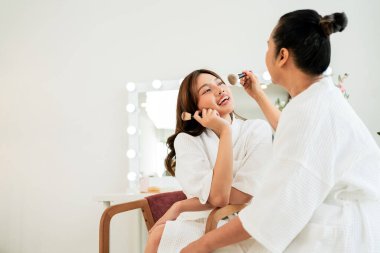 Beyaz spa elbiseli anne ve kızı, makyaj yaparken birlikte gülüyorlar. Parlak bir makyaj ortamında, sıcaklığı, kendine bakımı ve neşeli bağ kurma ideallerini yansıtıyorlar. Anneler Günü ve sağlık teması.