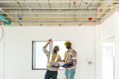 İki siyah mühendis bir inşaat sahasında tavan altyapısını inceliyor, biri dizüstü bilgisayarını tutarken yukarıya bakıyor. Resim takım çalışması, denetim ve modern yapı değerlendirmesini yakalar.