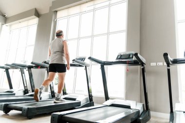 Yaşlı adam yağ kaybı, kalp sağlığı ve kilo kontrolü için parlak bir spor salonunda koşu bandında yürüyor. Sağlıklı günlük egzersiz ve kilo verme rutininin bir parçası olarak sağlık, sağlık ve aktif yaşam tarzını teşvik eder.