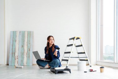 Yerde oturan genç bir kadın dizüstü bilgisayarı ve boya fırçasıyla DIY evini yenilemeyi planlıyor. Parlak modern odada aletler ve merdivenlerle çevrelenmiş halde tasarım yaratıcılığı ve üretkenliği dengeler..