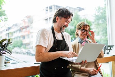 Gülümseyen kafe sahibi ve bayan danışman samimi kahve dükkanında dizüstü bilgisayarda birlikte çalışıyorlar. Bunlar iş büyümesi ve performansın artırılması için mali planlar ve pazarlama stratejileri üzerinde işbirliği yaparlar..