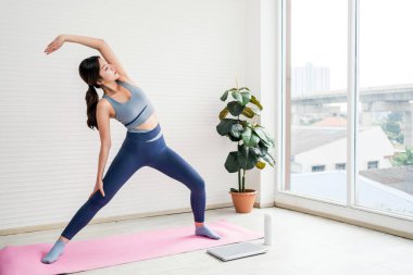 İçeride yoga yapan genç bir kadın, büyük pencereli parlak bir odada pembe bir paspasın üzerinde geriniyor. Sakinlik, odaklanmış enerji ve denge ile sağlık ve sağlıklı yaşam tarzını teşvik eder..
