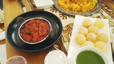 Etnik bir masa örtüsünde servis edilen pani puri, chutney, köri ve biryani gibi geleneksel yemeklerle yayılan canlı bir Hint aile yemeği kültürel sıcaklığı ve festival havasını çağrıştırıyor..