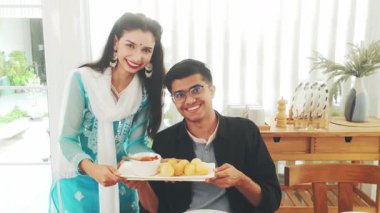 Mutlu Hintli çift geleneksel kıyafetleriyle gülümseyerek ve bir tabak pani puri sunarak neşeli bir ev kutlamasında kültürlerini, yemeklerini ve konukseverliklerini gururla sergiliyorlar..