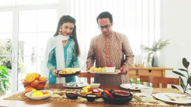 Geleneksel giyinmiş Hintli çift pani puri, köri, naan ve meyve gibi otantik yemeklerle bir aile yemeği masası hazırlıyorlar. Kültürü ve ev yapımı konukseverliği kutluyorlar..