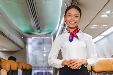 Gülümseyen uçuş görevlisi uçak koridorunda duruyor, uçuş hizmeti sunuyor. Ticari uçuşlarda konukseverlik, havayolu güvenliği ve yolcu bakımını temsil ediyor. Seyahat endüstrisi görselleri için ideal