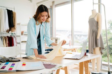 Modern bir stüdyoda çalışan genç bir kadın moda tasarımcısı dizüstü bilgisayardaki tasarımlardan bahsederken kumaş seçiyor. Küçük işletme sahibi çevrimiçi veya butik perakende pazarları için özel kıyafetler üretiyor.