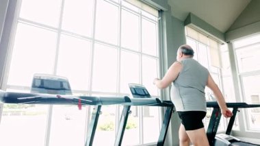 Spor salonunda koşu bandında yürüyen kilolu son sınıf öğrencisi daha iyi bir sağlık ve uzun yaşam için zinde olmayı kucaklıyor. Doğal ışık, sağlık, egzersiz ve aktif yaşlanmayı teşvik eden modern kapalı spor salonu..
