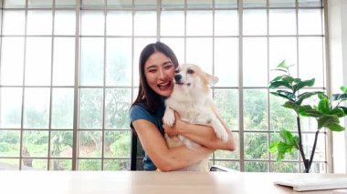 Evde elinde sevimli bir Corgi köpeği olan neşeli genç bir kadın gülümseyerek kameraya bakıyor, büyük bir pencerenin önünde oturuyor, neşeyi, kaynaşmayı ve evcil hayvan dostu bir yaşam tarzını temsil ediyor..