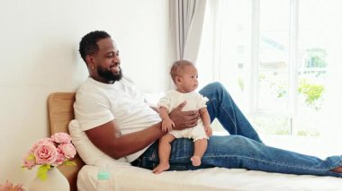 Sevgi dolu bir baba, parlak, rahat bir yatak odasında bebeğini kucağında tutar. Otantik, modern ebeveynliği sembolize eder. Babalık neşesi ve farklı bir aile ortamında günlük bağları sembolize eder..