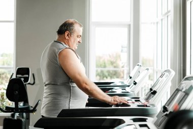 Parlak bir spor salonunda koşu bandı kullanmaya hazırlanan kilolu yaşlı bir adam sağlığını ve sağlıklı bir yaşam tarzını destekliyor. Fitness, sağlıklı yaşlanma ve kişisel refahı geliştirmeye odaklanmış..