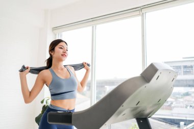 Kendine güvenen genç bir kadın koşu bandında egzersiz yapıyor, havluyu tutarken antrenman sonrası gülümsüyor. Parlak bir spor salonu ortamında sağlıklı bir yaşam tarzı, zindelik motivasyonu ve sağlığını yakalıyor..